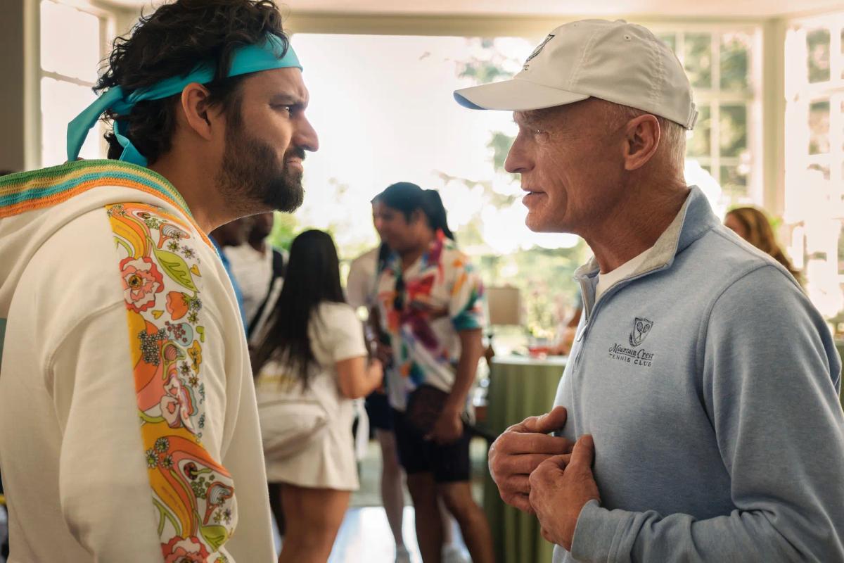 Dusty Boyd (Jake Johnson) in a floral robe and teal headband talks to his father Chuck Boyd (Ed Harris), who wears a club quarter-zip and cap.