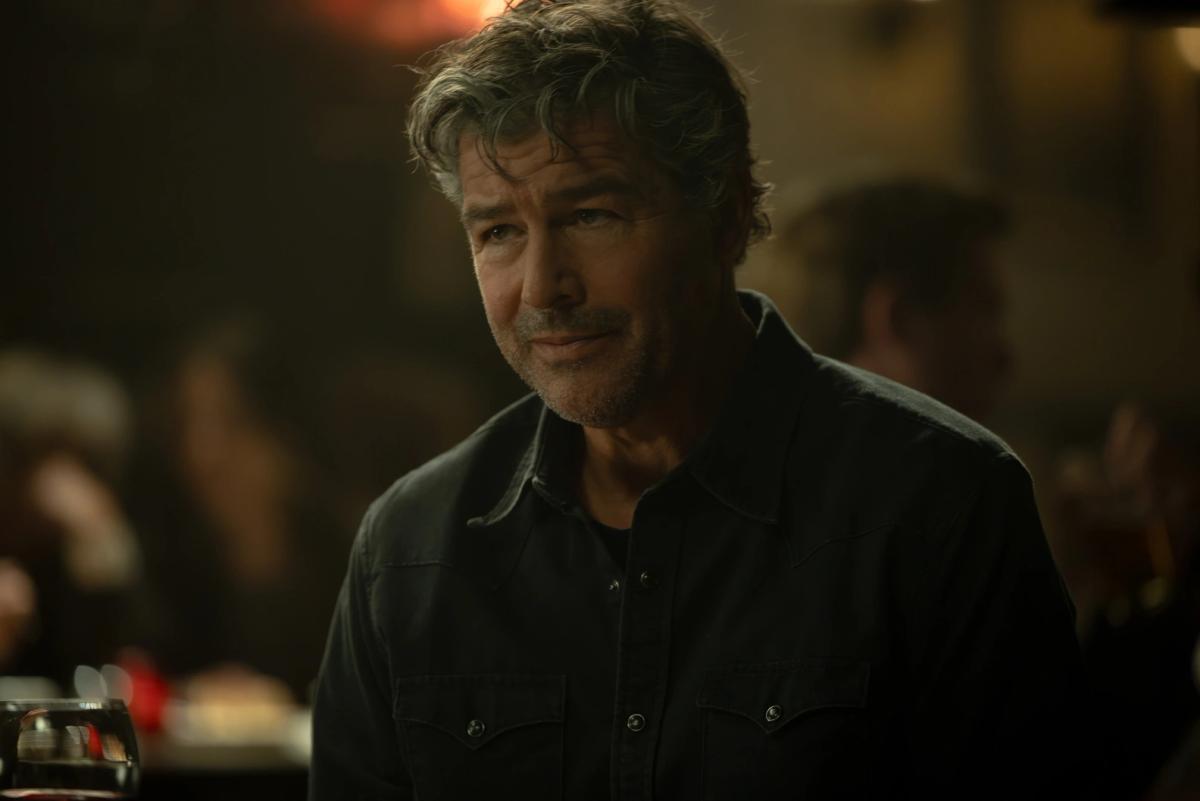 Kyle Chandler as Hal Jordan, grey-haired and half smiling, in a dark shirt at a busy restaurant.