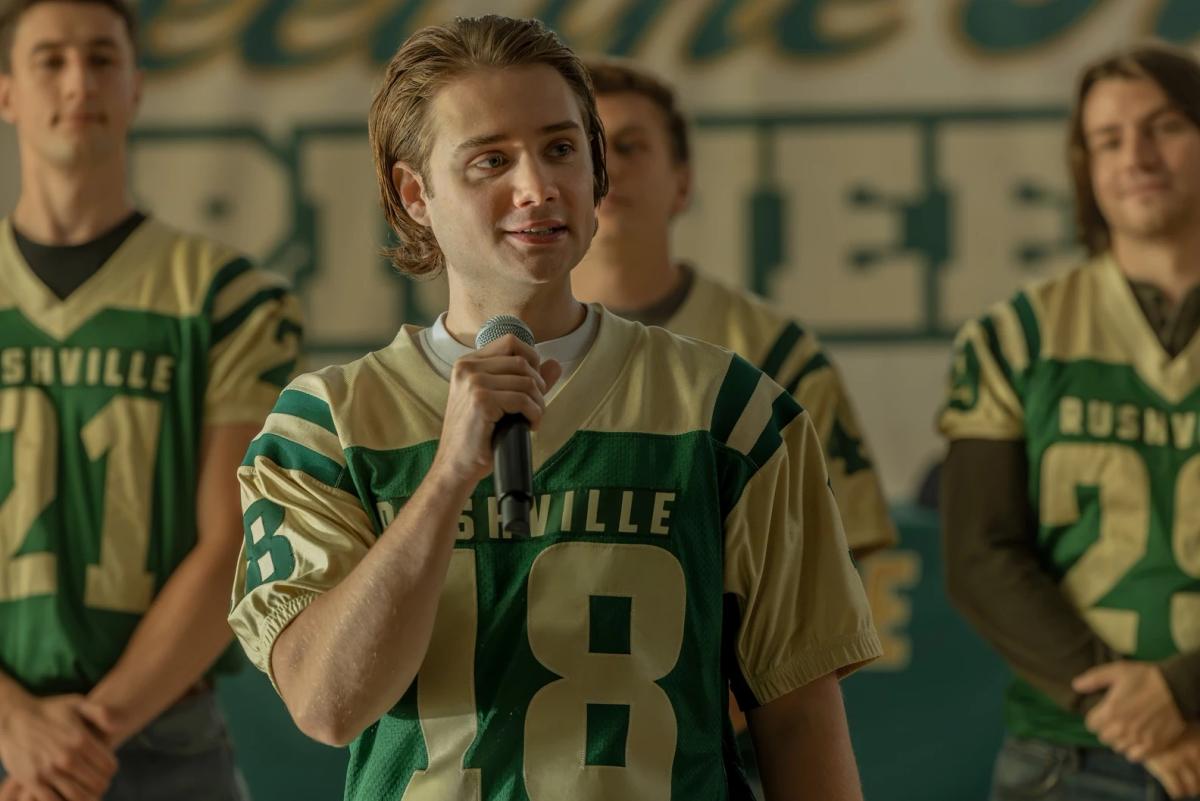 Shea Pritchard as Noah Macon in a green and gold Rushville number 18 jersey, speaking into a microphone in the school gym with teammates behind him.