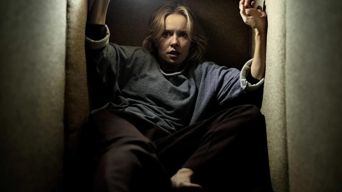 Gemma (Amelia Eve) wedged in a narrow gap between two sofa cushions, arms braced against the walls above her, lit from below in Insidious: Out of the Further.