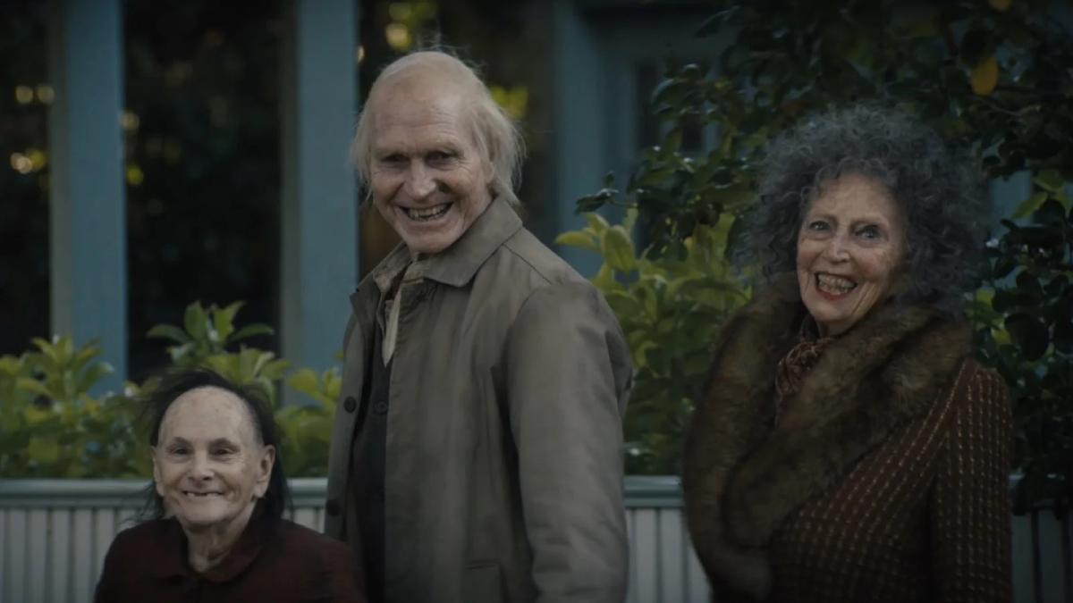 Three pale, decrepit elderly figures — a bald man in a raincoat flanked by two women — standing outside a suburban house and smiling directly at the camera in Insidious: Out of the Further.
