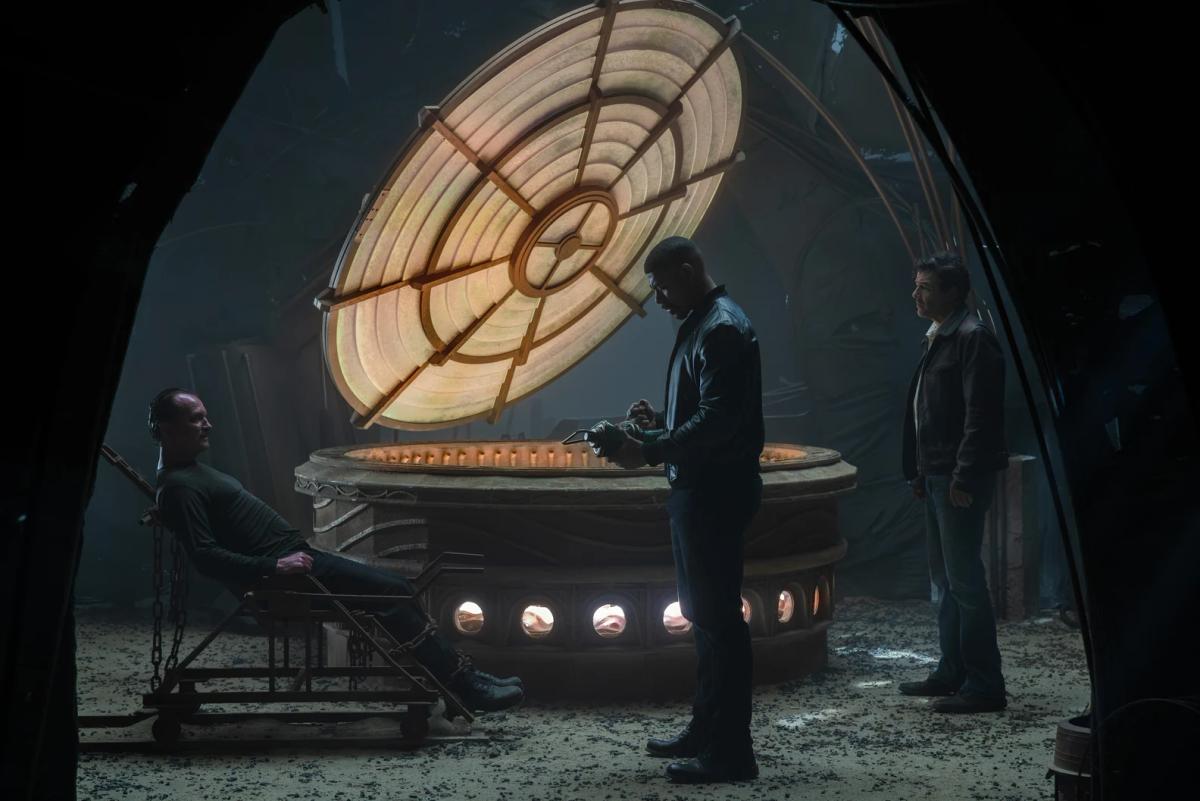 Ulrich Thomsen as Sinestro chained to a chair beside a large glowing forge, while Aaron Pierre as John Stewart holds a lantern and Kyle Chandler as the Halogram looks on.