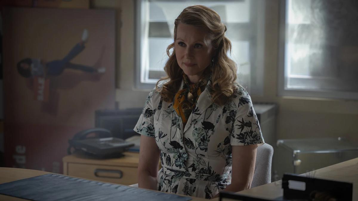 Laura Linney as a Guardian of the Universe in human form, seated behind a desk in a bare office.