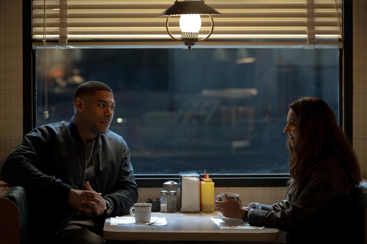 Aaron Pierre as John Stewart and Poorna Jagannathan as Zoe Macon sitting opposite each other in a lit diner booth at night.