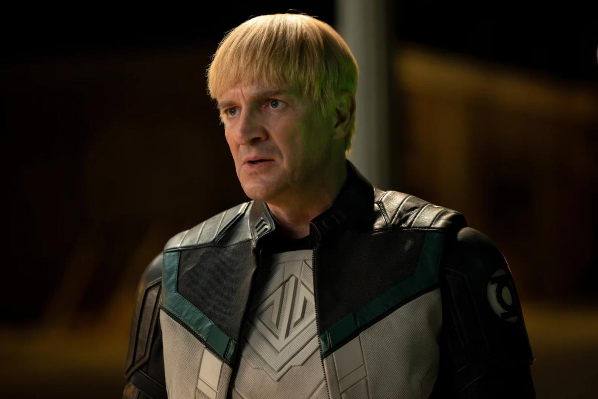 Nathan Fillion as Guy Gardner, with a blond bowl haircut, in a black and white Green Lantern uniform with the Corps emblem on his shoulder.