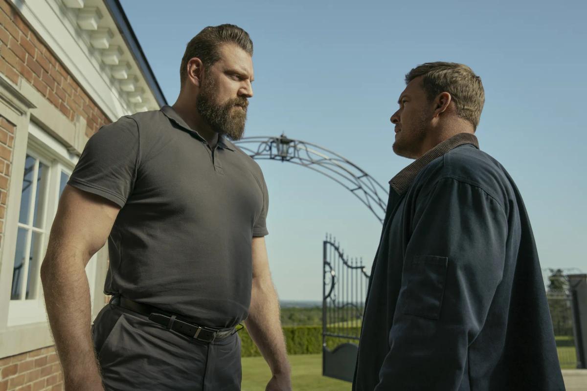 Olivier Richters as Paulie, bearded and in a grey polo shirt, looks down at Alan Ritchson as Jack Reacher outside a brick house, with an iron gate and a clear sky behind them.
