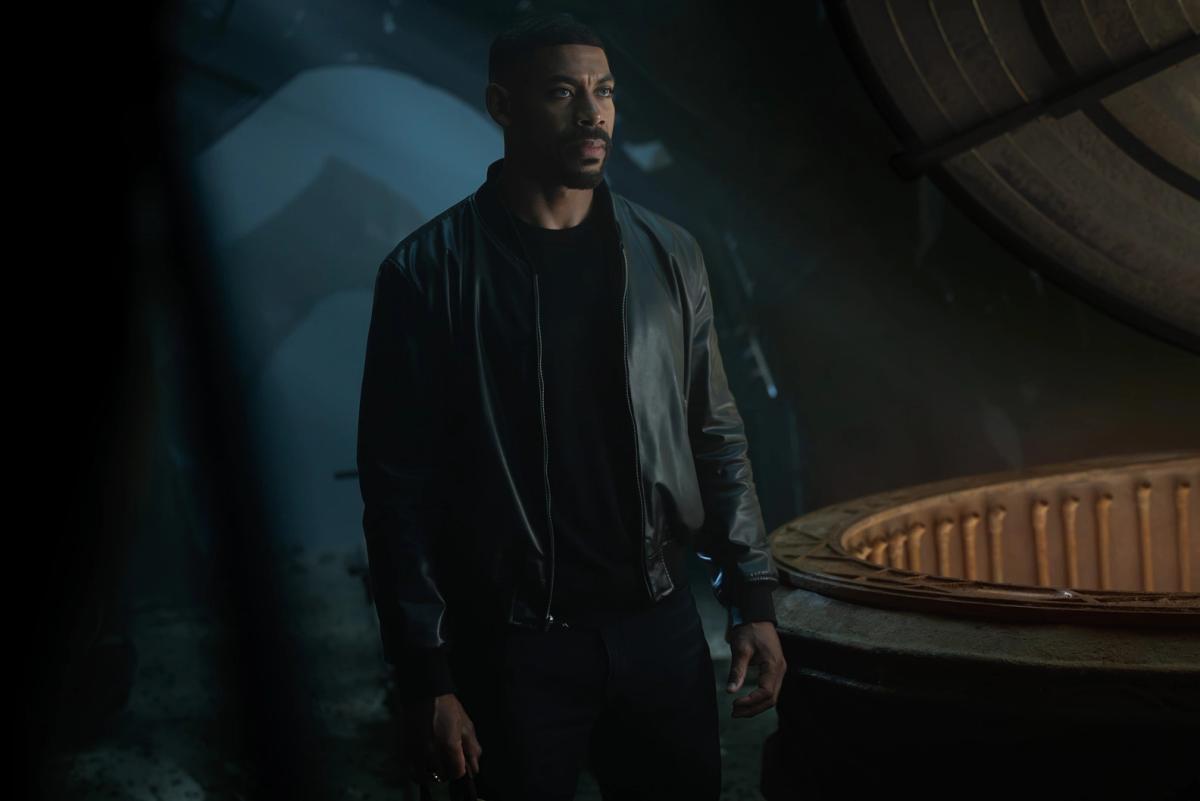 Aaron Pierre as John Stewart in a black leather jacket, standing in a dark chamber beside a huge circular structure that glows amber inside.