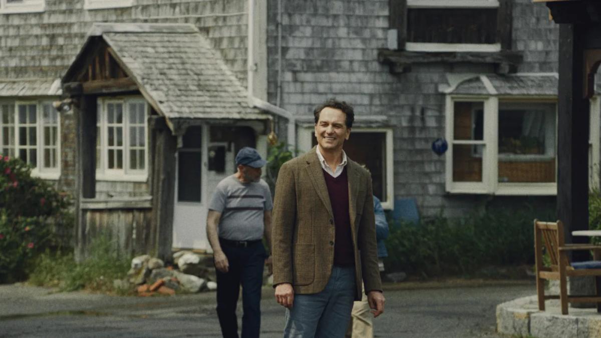 Matthew Rhys as Tom Loftis walks through the shingled centre of Widow's Bay in a tweed jacket, smiling, while a resident watches him from behind.