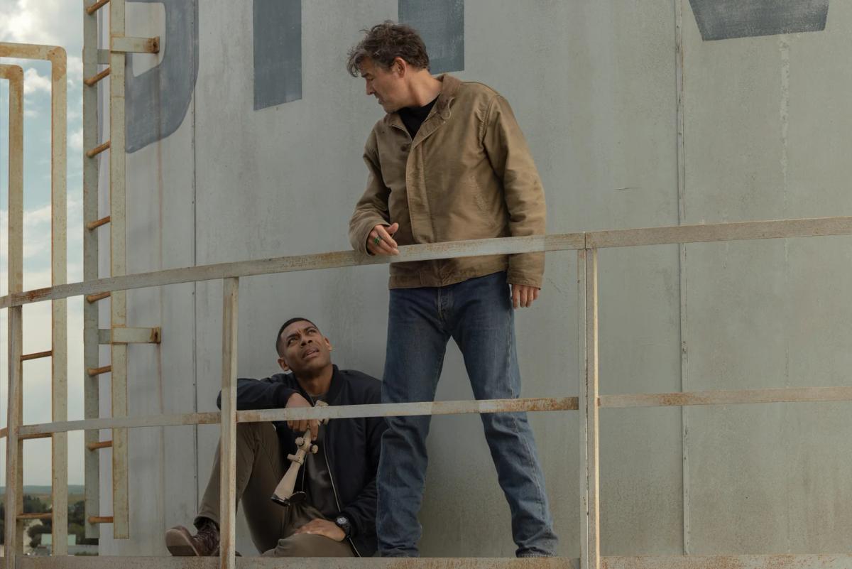 Kyle Chandler as Hal Jordan standing over Aaron Pierre as John Stewart, who is sitting on a grain silo walkway with a telescope.