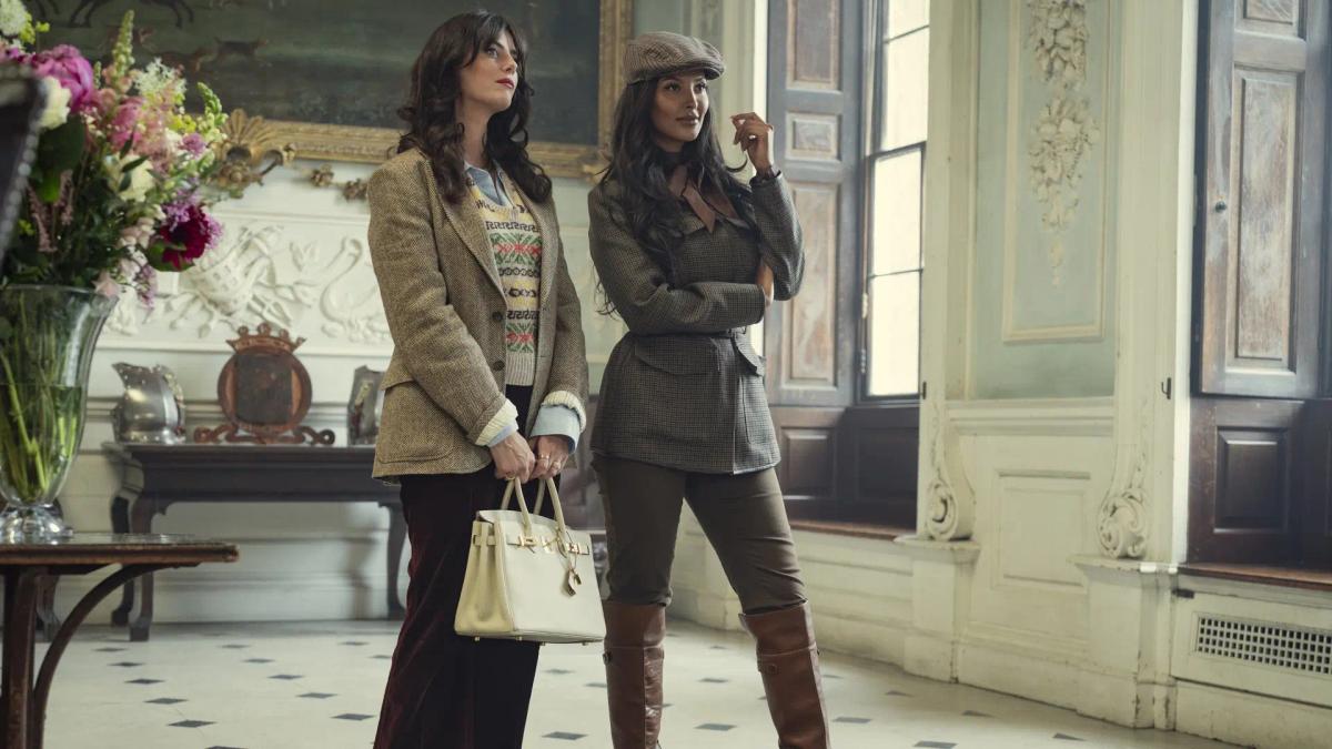 Kaya Scodelario as Susie Glass and Maya Jama as Aisha standing in the hall at Halstead in tweed shooting clothes.