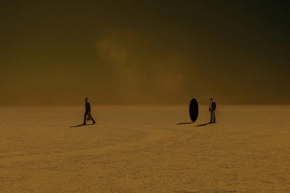 Aaron Pierre as John Stewart, carrying a backpack, and Kyle Chandler as the Halogram on a vast cracked yellow desert under a dark sky, with a black oval portal standing between them.