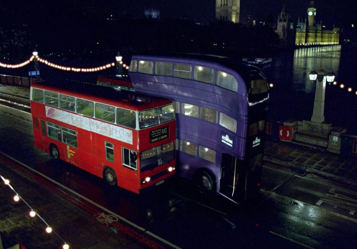 The purple triple-decker Knight Bus squeezing past a red London bus on a wet bridge at night, with the lit Houses of Parliament behind it.