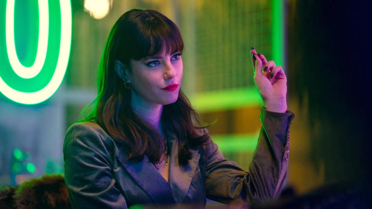 Kaya Scodelario as Susie Glass, lit by green neon, raising a hand mid-conversation.