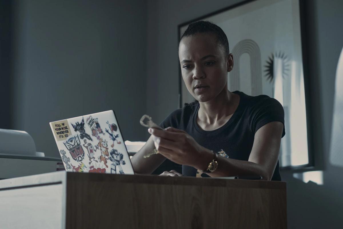 Maria Sten as Frances Neagley sits at a desk in a dim room, working at a sticker-covered laptop and turning a small metal object over in her fingers.