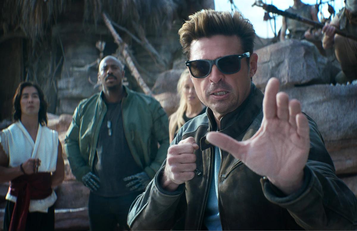 Karl Urban as Johnny Cage in sunglasses and a leather jacket, raising his hands in a fighting pose, with Ludi Lin as Liu Kang, Mehcad Brooks as Jax and Jessica McNamee as Sonya Blade behind him.