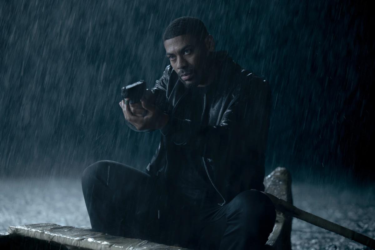Aaron Pierre as John Stewart crouched in a small boat in heavy rain at night, holding a handgun.
