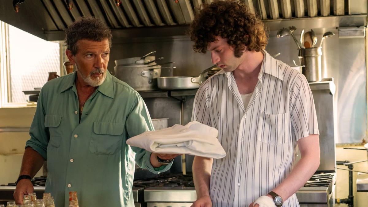 Antonio Banderas as Chef holding out a folded set of kitchen whites to Dominic Sessa as Tony Bourdain, whose hand is bandaged.