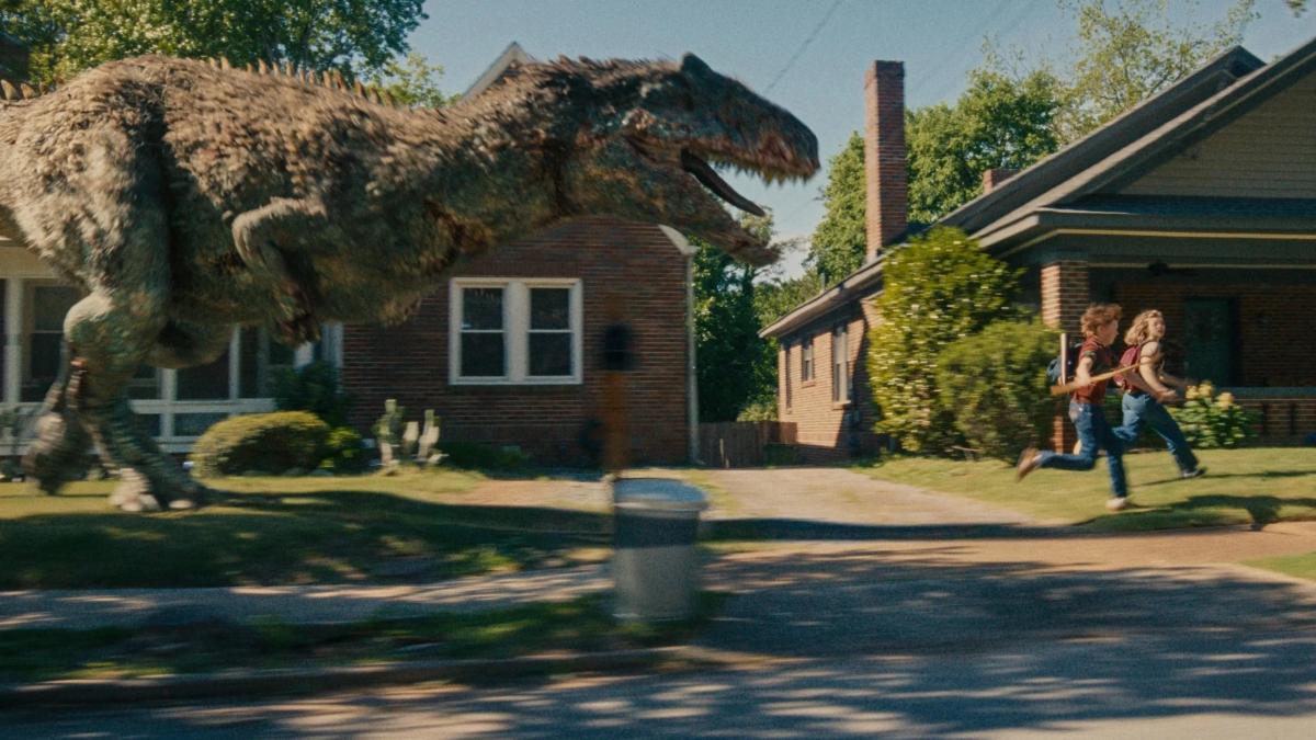 A large feathered predator running past suburban houses in daylight as two children flee across a lawn.