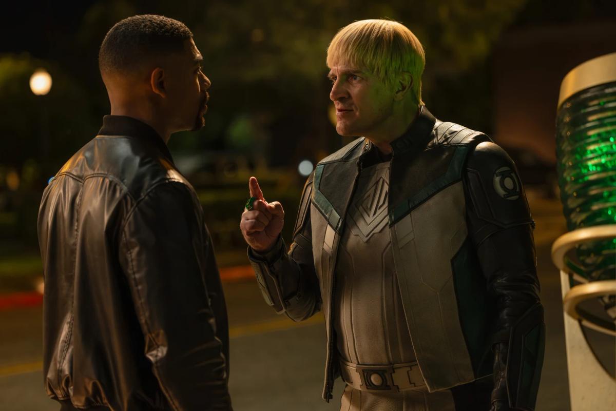 Nathan Fillion as Guy Gardner, in a white and black Green Lantern suit, pointing a finger at Aaron Pierre as John Stewart at night beside a glowing green beacon.