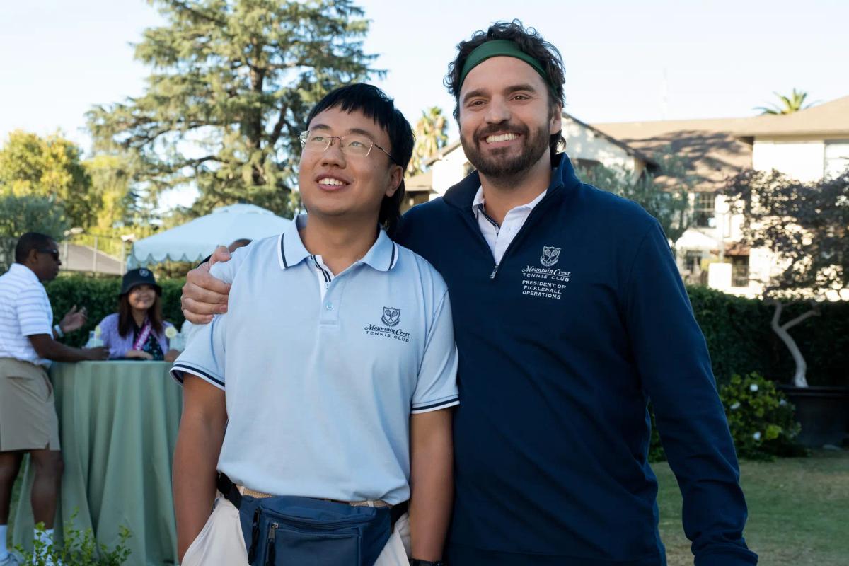 PJ (Aaron Chen) in a club polo stands arm in arm with Dusty Boyd (Jake Johnson) in a navy club quarter-zip, outdoors at the tennis club.