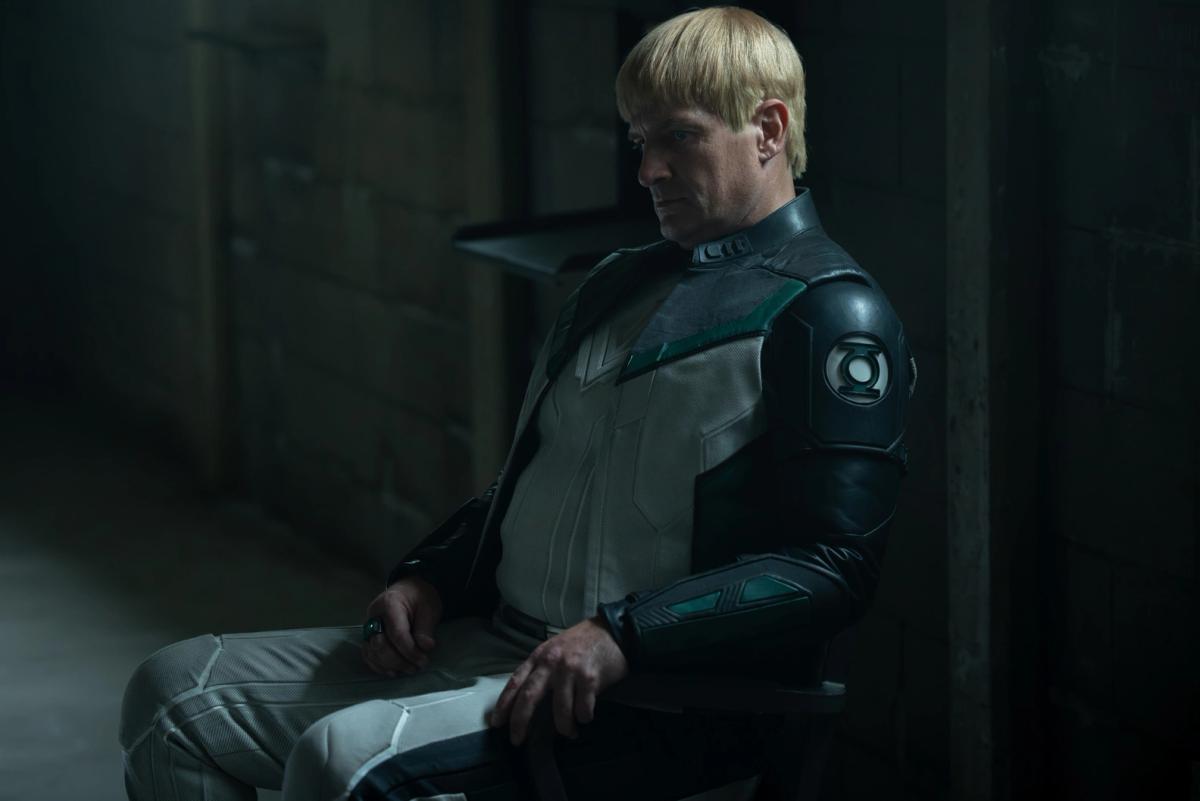 Nathan Fillion as Guy Gardner in Lanterns, seated in a dim room in a Green Lantern uniform with a green ring on his hand.