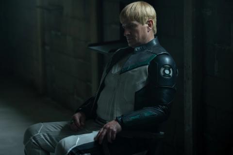 Nathan Fillion as Guy Gardner in Lanterns, seated in a dim room in a Green Lantern uniform with a green ring on his hand.