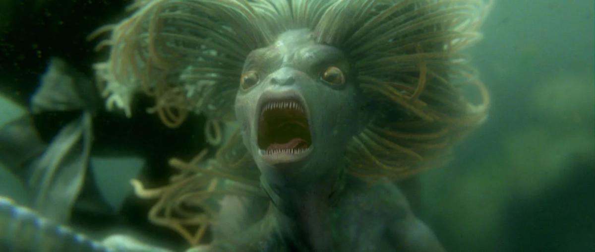 A merperson with pale skin and wild hair bares its teeth underwater in the Black Lake.