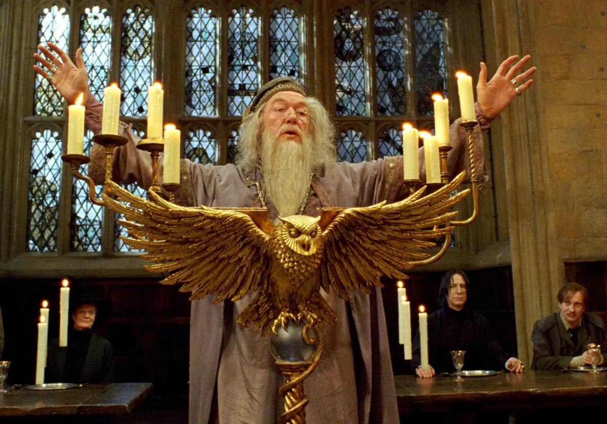 Michael Gambon as Albus Dumbledore addressing the Great Hall with both arms raised, watched from the staff table by Maggie Smith as Minerva McGonagall, Alan Rickman as Severus Snape and David Thewlis as Remus Lupin.