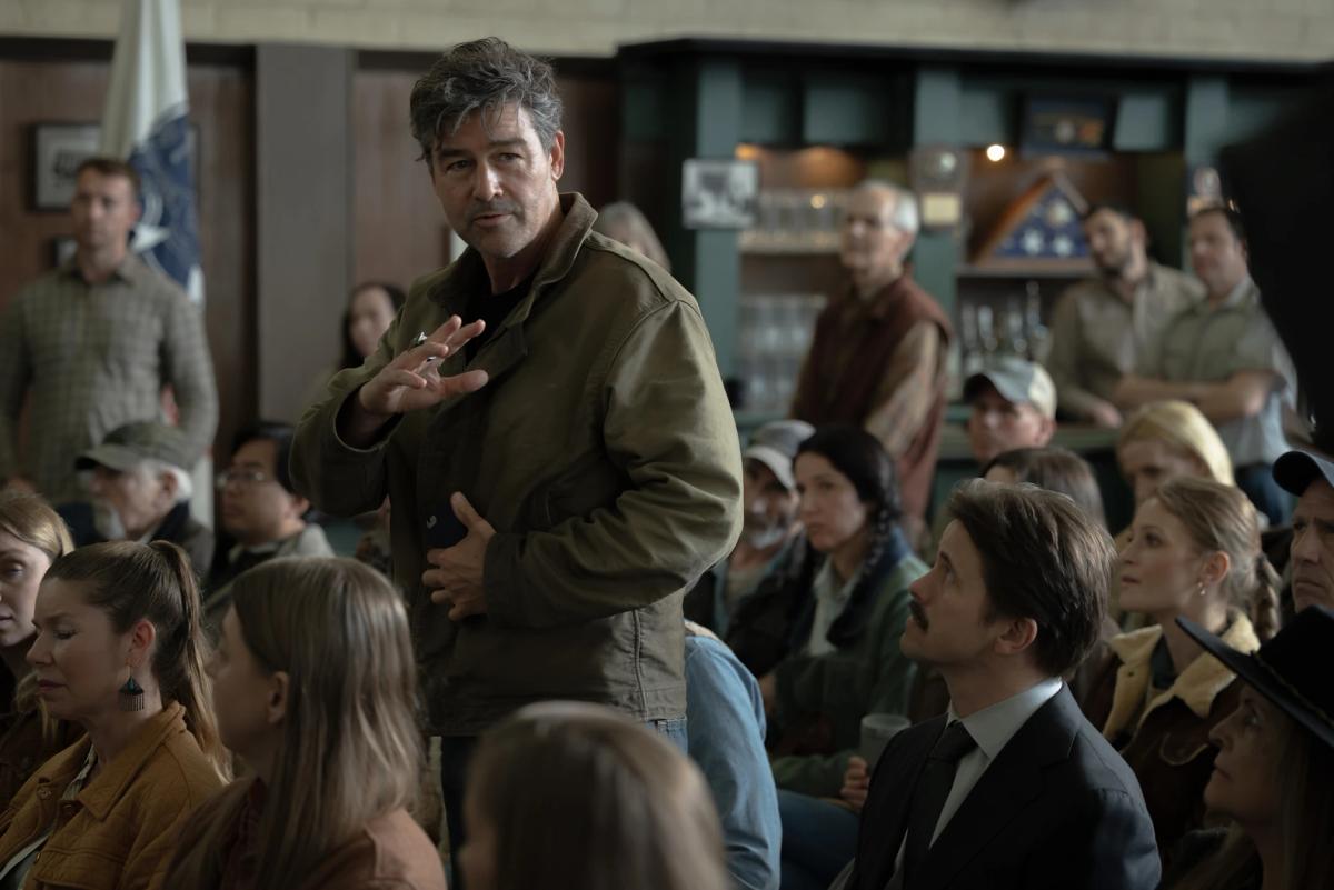 Kyle Chandler as Hal Jordan standing to speak in a crowded town hall, surrounded by seated Rushville residents, with Jason Ritter as Billy Macon looking up at him.