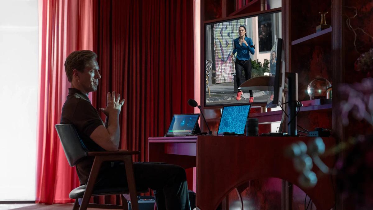The Caller sits at a desk in a red-lit room, watching Maia run on a large screen beside a map and a glucose readout.