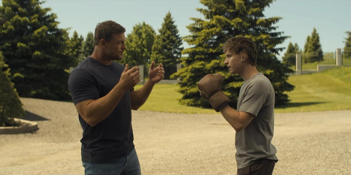 Alan Ritchson as Jack Reacher coaches Johnny Berchtold as Richard Beck, who has his guard up in a pair of old brown leather boxing gloves, on a gravel drive lined with evergreen trees.