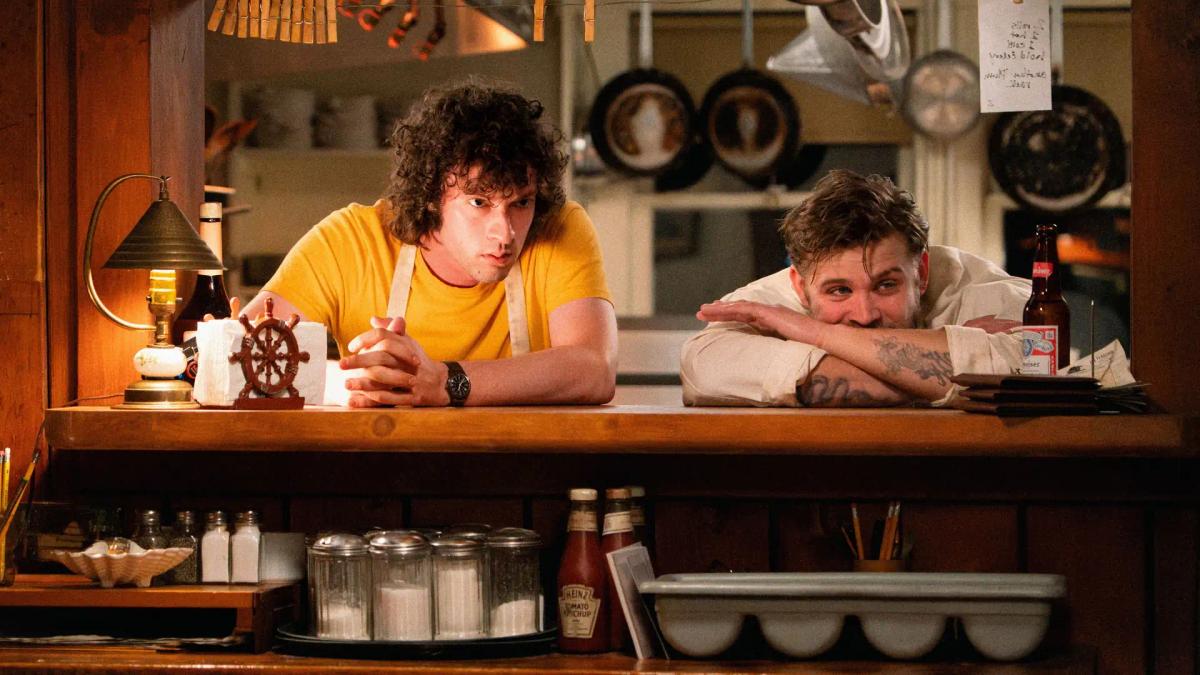 Dominic Sessa as Tony Bourdain leaning on the pass beside Leo Woodall as Sal, who has his head down on his folded arms next to a beer.