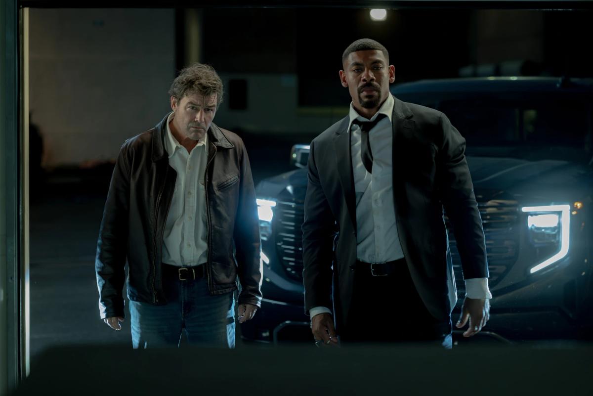 Kyle Chandler as the Halogram, in a brown leather jacket, beside Aaron Pierre as John Stewart, in a dark suit with his tie loosened, at night in front of a pickup truck's headlights.
