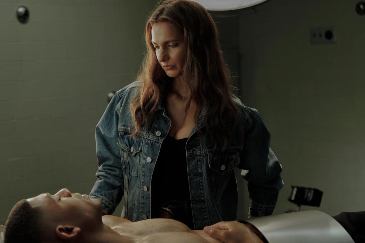 Poorna Jagannathan as Zoe Macon standing over Aaron Pierre as John Stewart, who lies on a hospital table.