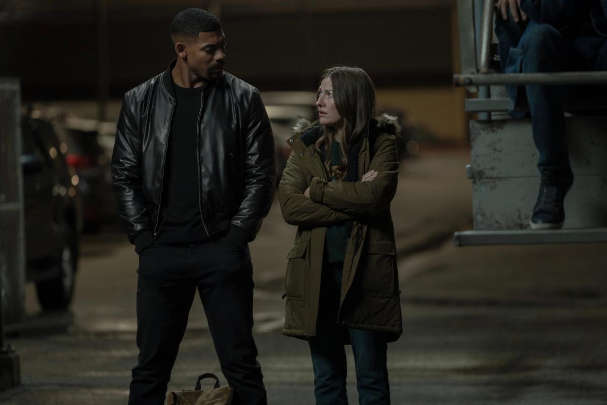 Aaron Pierre as John Stewart and Kelly Macdonald as Kerry Kane, arms folded inside a parka, standing together at night beside the stands.