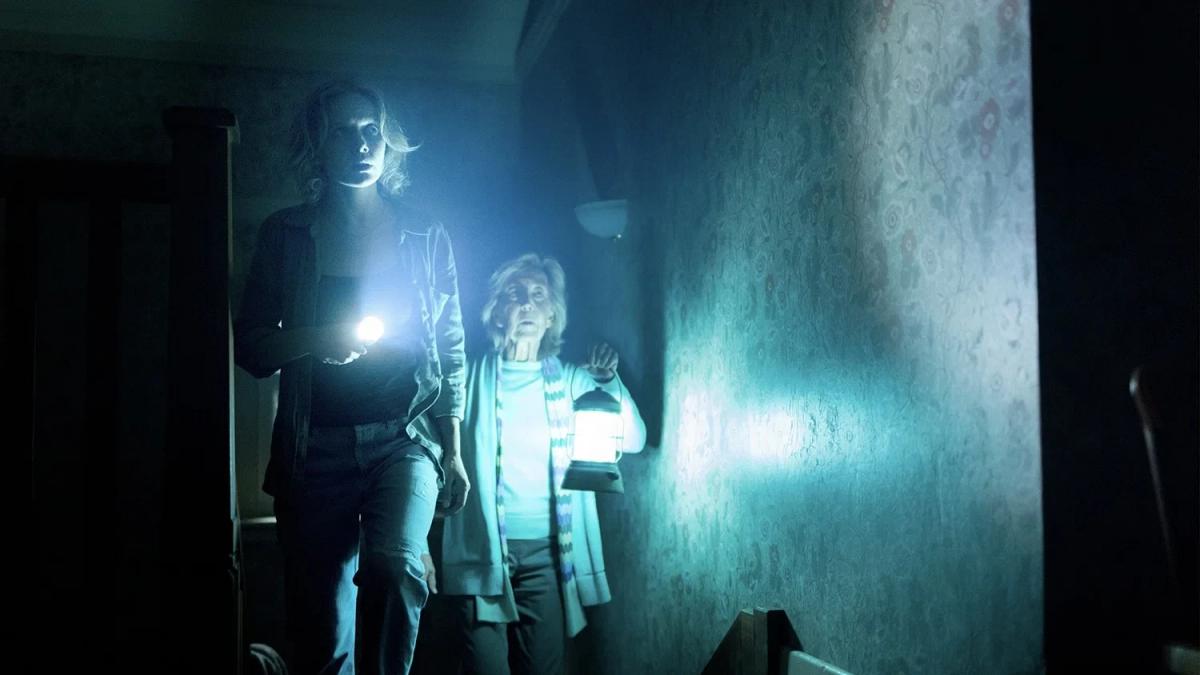 Gemma (Amelia Eve) holds a torch as she moves through a dark hallway, followed by Elise Rainier (Lin Shaye) carrying a lit lantern, in Insidious: Out of the Further.