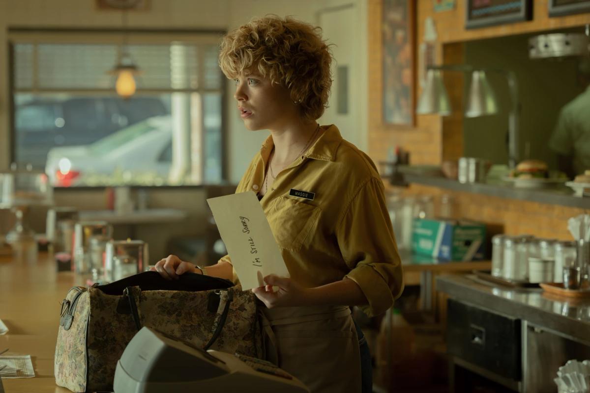 Hannah Christine Shetler as Maggie behind the café counter in her yellow uniform, holding an envelope marked "I'm still sorry" beside an open handbag.