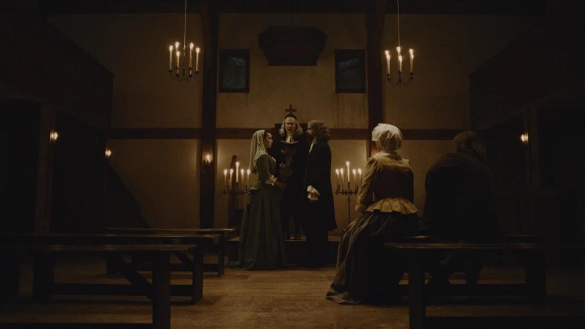 A candlelit meeting house in 1702: Betty Gilpin as Sarah Westcott, veiled and in a green gown, marries Hamish Linklater as Richard Warren before a minister, watched by two seated witnesses.