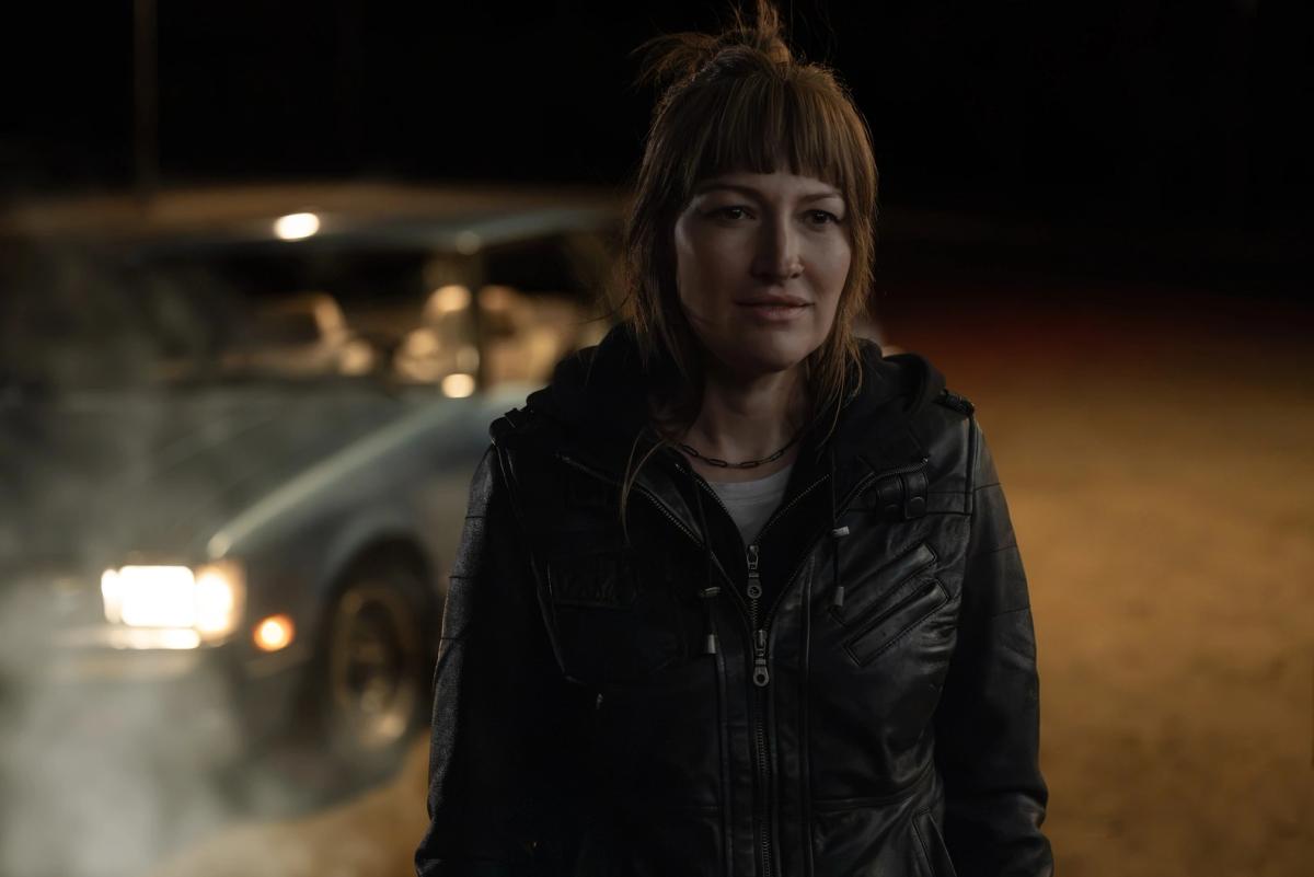 Kelly Macdonald as a younger Kerry Kane standing at night in a leather jacket, car headlights behind her.