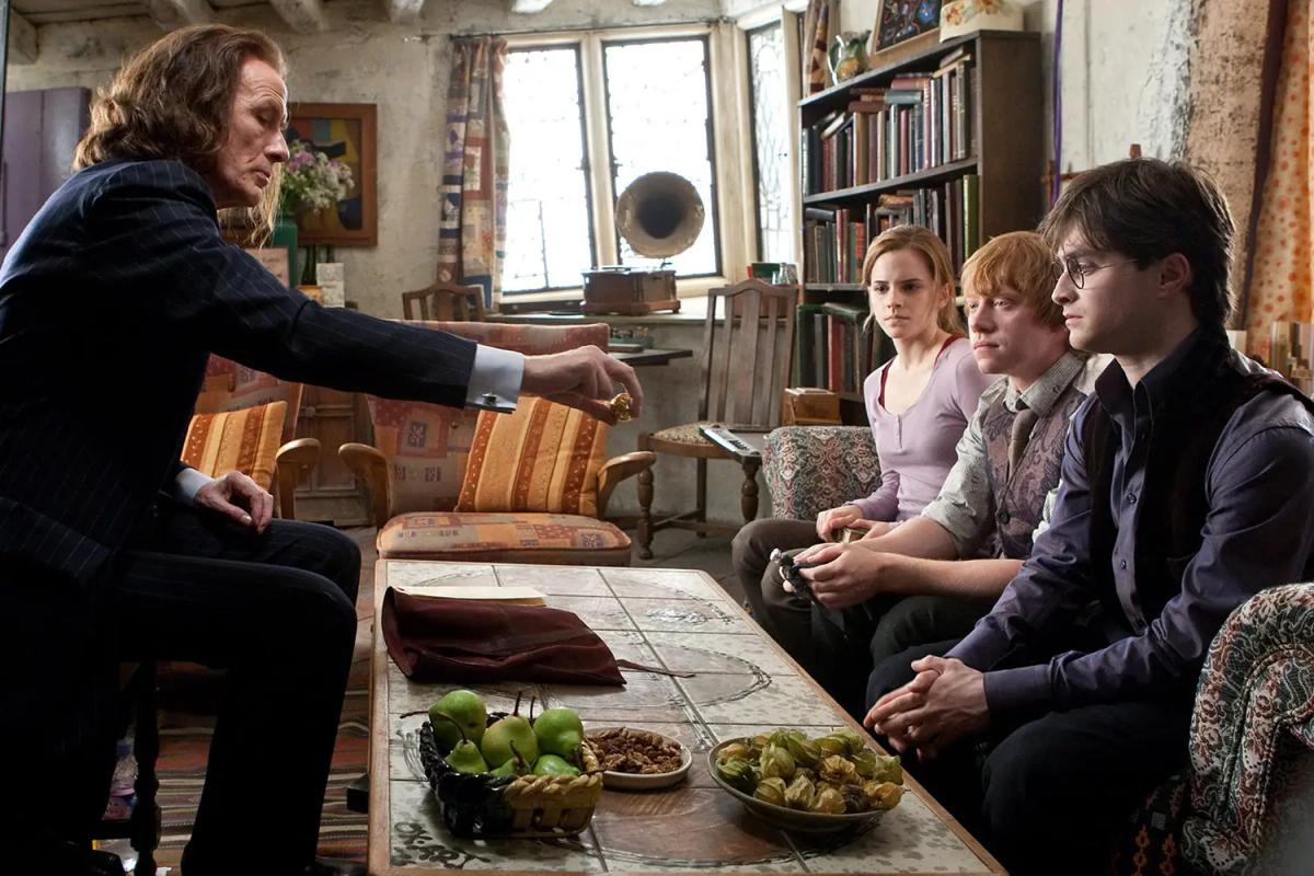Rufus Scrimgeour (Bill Nighy) leans across a cluttered table to hand something to Harry Potter (Daniel Radcliffe), with Ron Weasley (Rupert Grint) and Hermione Granger (Emma Watson) seated beside him.