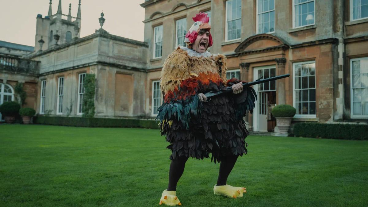 Daniel Ings as Freddy Horniman, in a full chicken costume on the lawn at Halstead, holding a shotgun and shouting.
