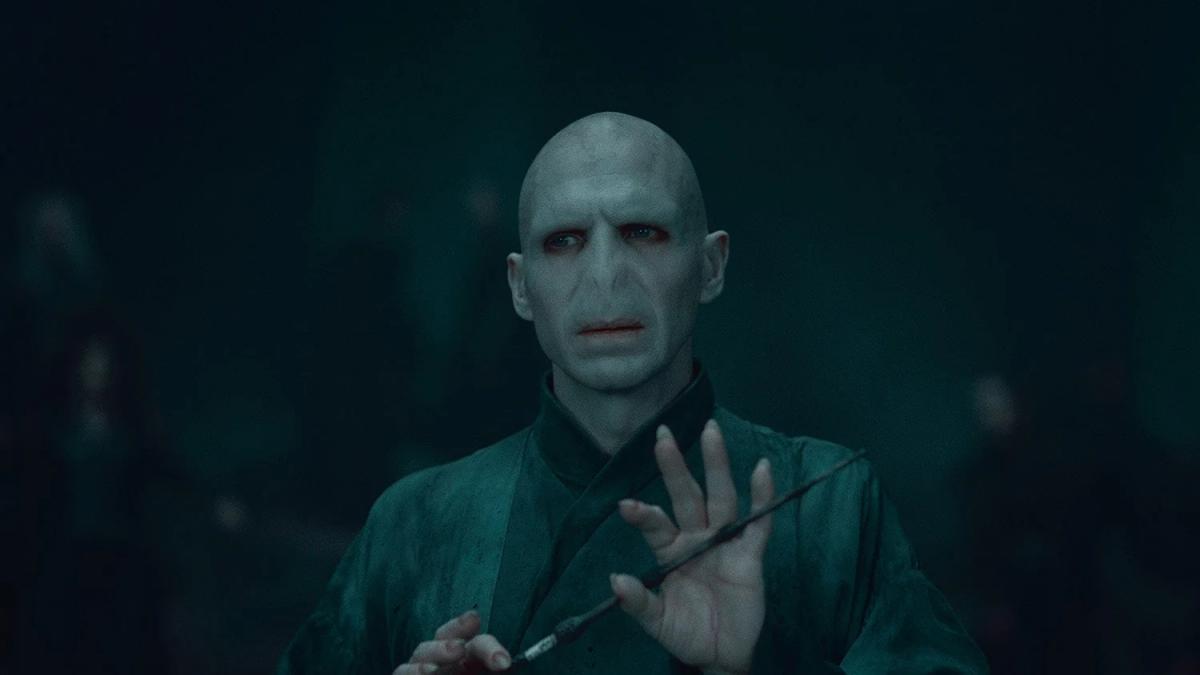 Ralph Fiennes as Lord Voldemort holding the Elder Wand up in front of his face, his hand open beside it.
