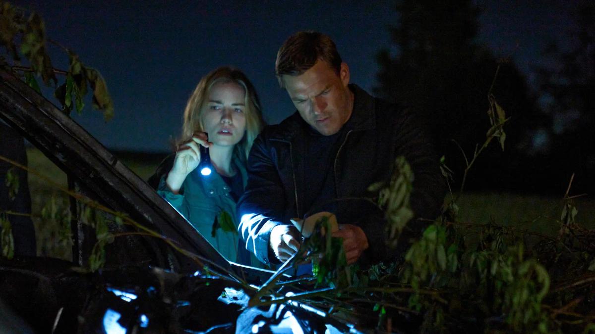 Willa Fitzgerald as Roscoe Conklin holds a torch while Alan Ritchson as Jack Reacher searches a wrecked car hidden under branches at night.