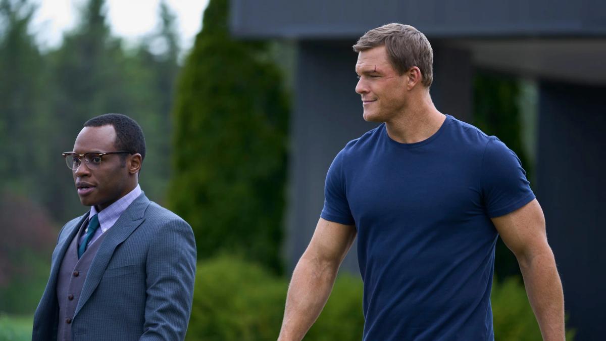Alan Ritchson as Jack Reacher walks outdoors beside Malcolm Goodwin as Detective Oscar Finlay, a head taller and roughly twice as broad.