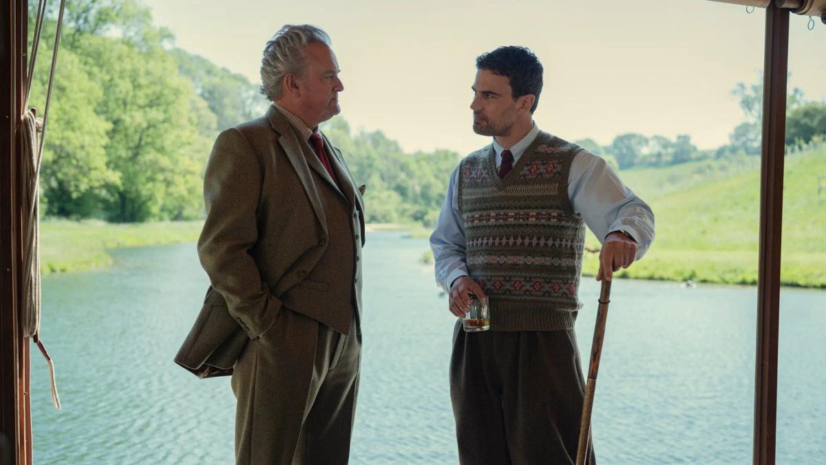 Hugh Bonneville as Lord Hawthorne talking with Theo James as Eddie Horniman in a boathouse looking out over a lake.