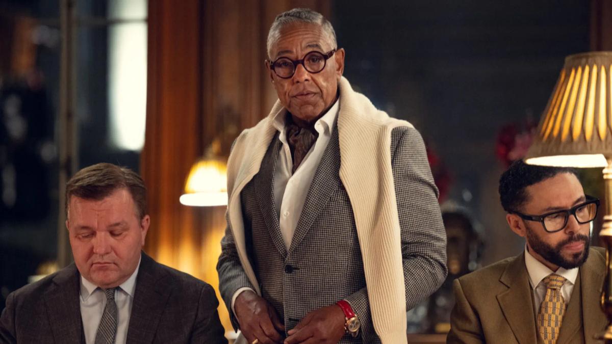 Giancarlo Esposito as Stanley Johnston, standing over two seated associates in a panelled drawing room.