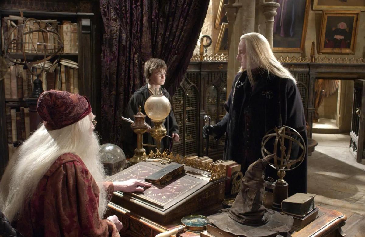 Lucius Malfoy (Jason Isaacs) stands across the desk from Albus Dumbledore (Richard Harris) in the headmaster's office, with Harry Potter (Daniel Radcliffe) behind them and the Sorting Hat on the desk.