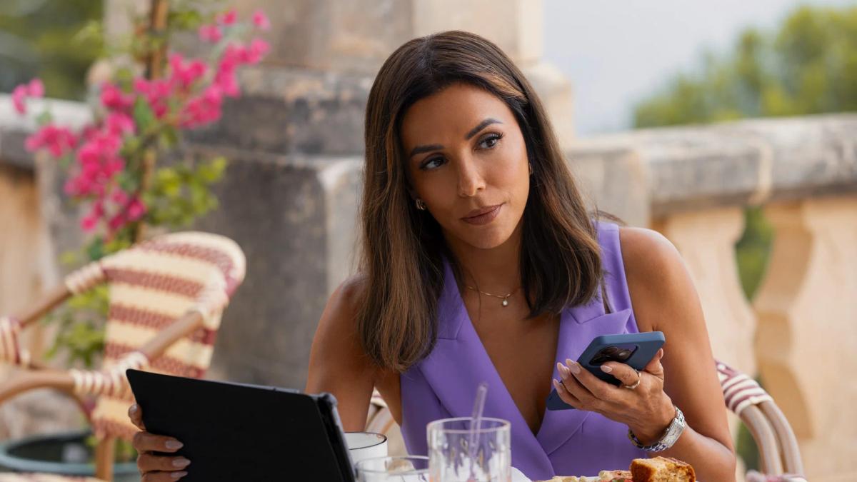 Eva Longoria as Isolde Pera holding a tablet in one hand and a phone in the other at an outdoor table.