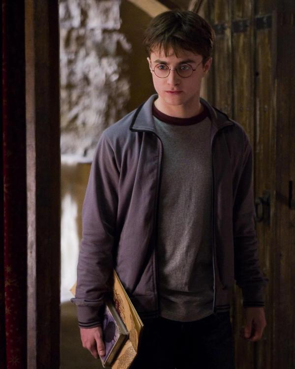 Daniel Radcliffe as Harry Potter walks through a doorway carrying his copy of Advanced Potion-Making.