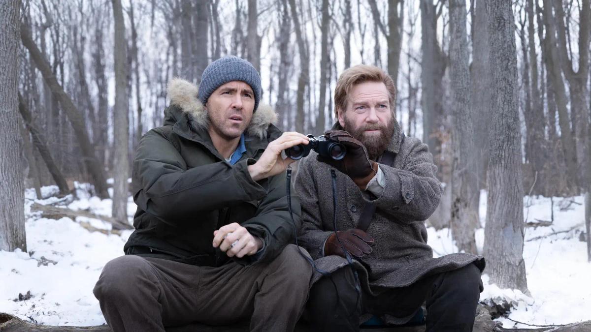 Ryan Reynolds as Troy Kelly and Kenneth Branagh as Nikolai Ustinov sharing a pair of binoculars on a log in a snowy forest